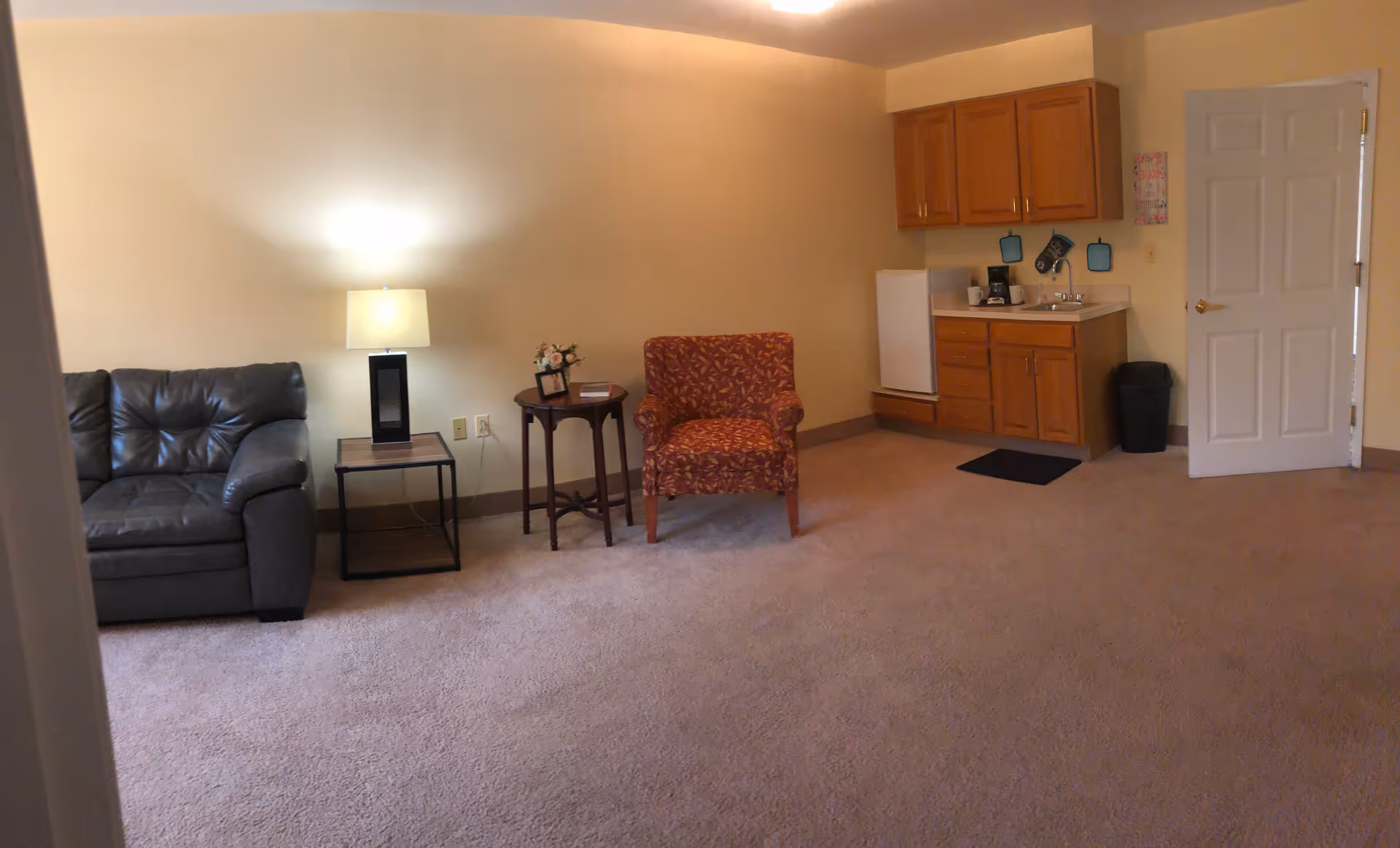 A cozy living area with a dark leather couch on the left, a small side table with a lamp, a round wooden table with flowers and a picture frame, and a patterned armchair. On the right side, there is a small kitchenette with wooden cabinets, a mini refrigerator, a coffee maker, and a sink. A white door is visible next to the kitchenette. The room has beige walls and carpeted flooring.