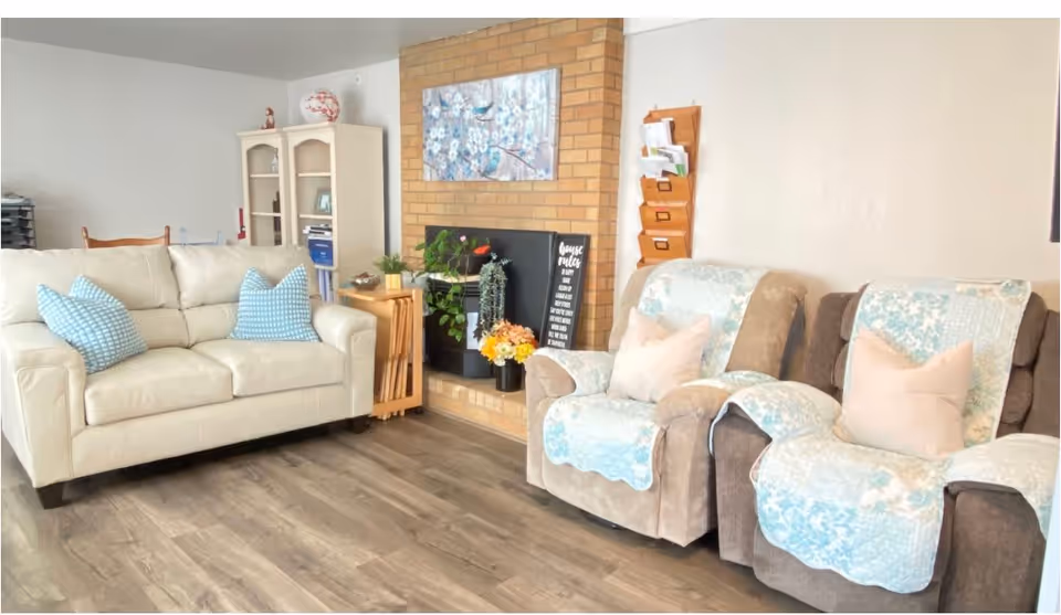 Cozy living room with a cream loveseat, two upholstered recliners draped with blue patterned throws, a brick fireplace and shelving.