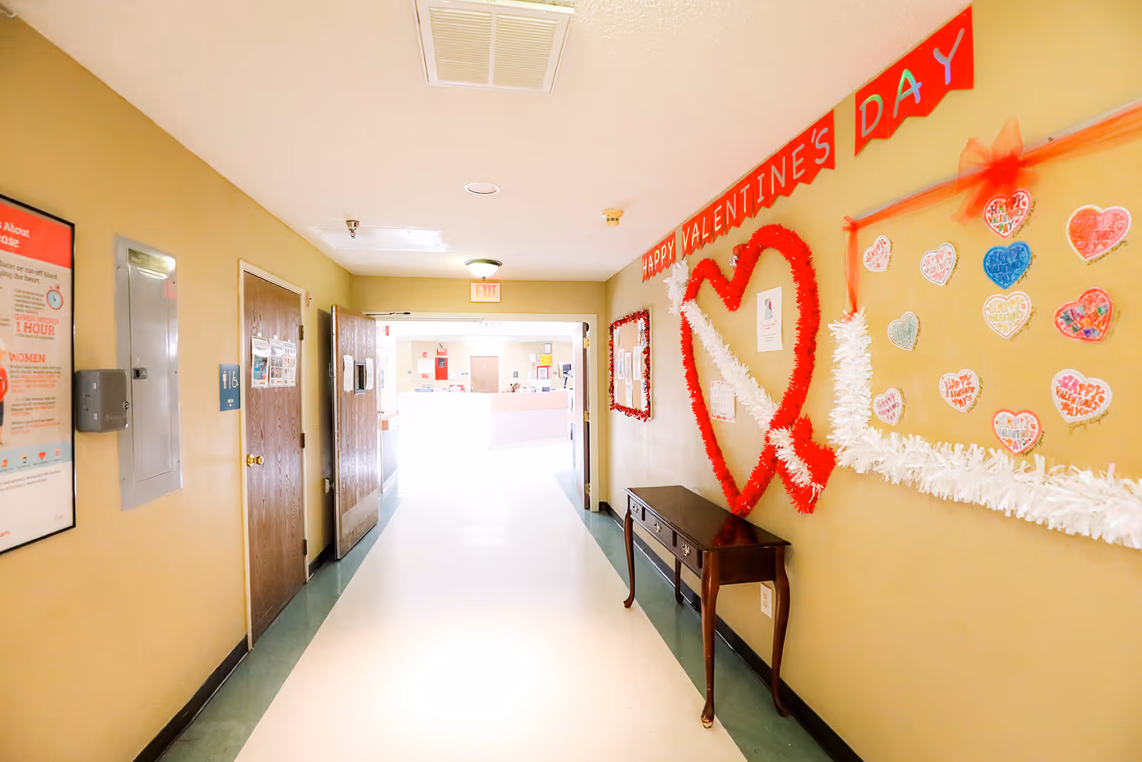 Interior hallway decorated for Valentine's Day with heart garlands, a bulletin board, doors, and a small table.