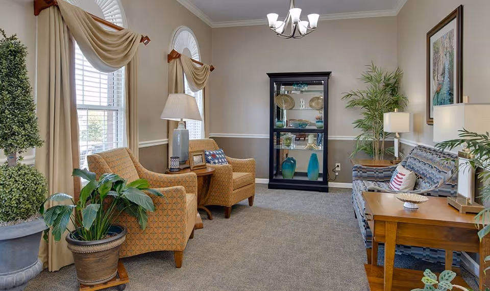A cozy living room area with two patterned armchairs and a sofa, all arranged around wooden side tables with lamps. The room features large windows with beige curtains, several potted plants, a black display cabinet with decorative items, and a framed painting on the wall. The carpet is light gray, and the walls are painted beige with white trim.