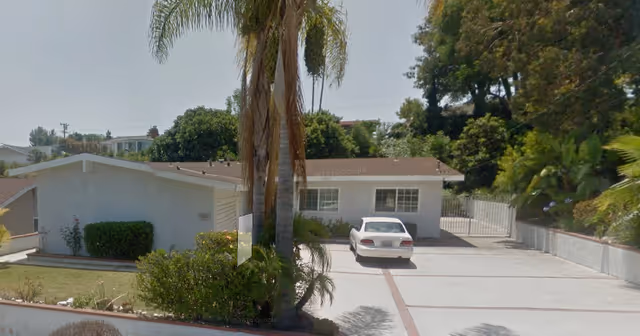 Exterior view of a single-story residential building with a white car parked in the driveway. The building is surrounded by trees and bushes, with a palm tree prominently in the foreground.