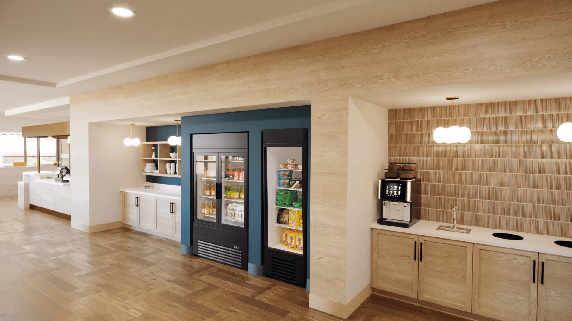 Interior view of a modern kitchen area with wooden flooring and cabinetry. There are two glass-door refrigerators stocked with beverages and snacks, a coffee machine on the counter, a small sink, and pendant lights hanging from the ceiling. The walls have a combination of wood paneling and blue paint accents.