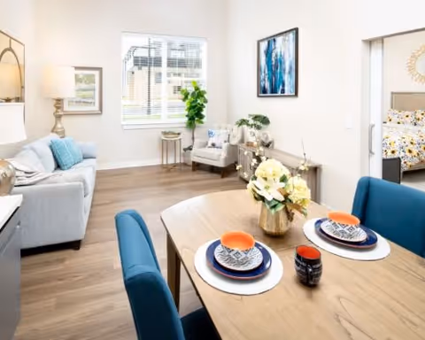 Bright open-plan living and dining area with a set table in the foreground, sofa and armchair by a window, and a glimpse of a bedroom.