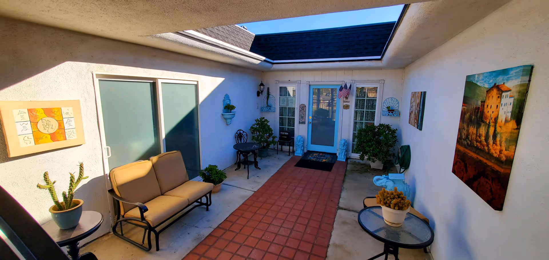 Sunlit courtyard patio with seating, potted plants, and wall art near glass doors.