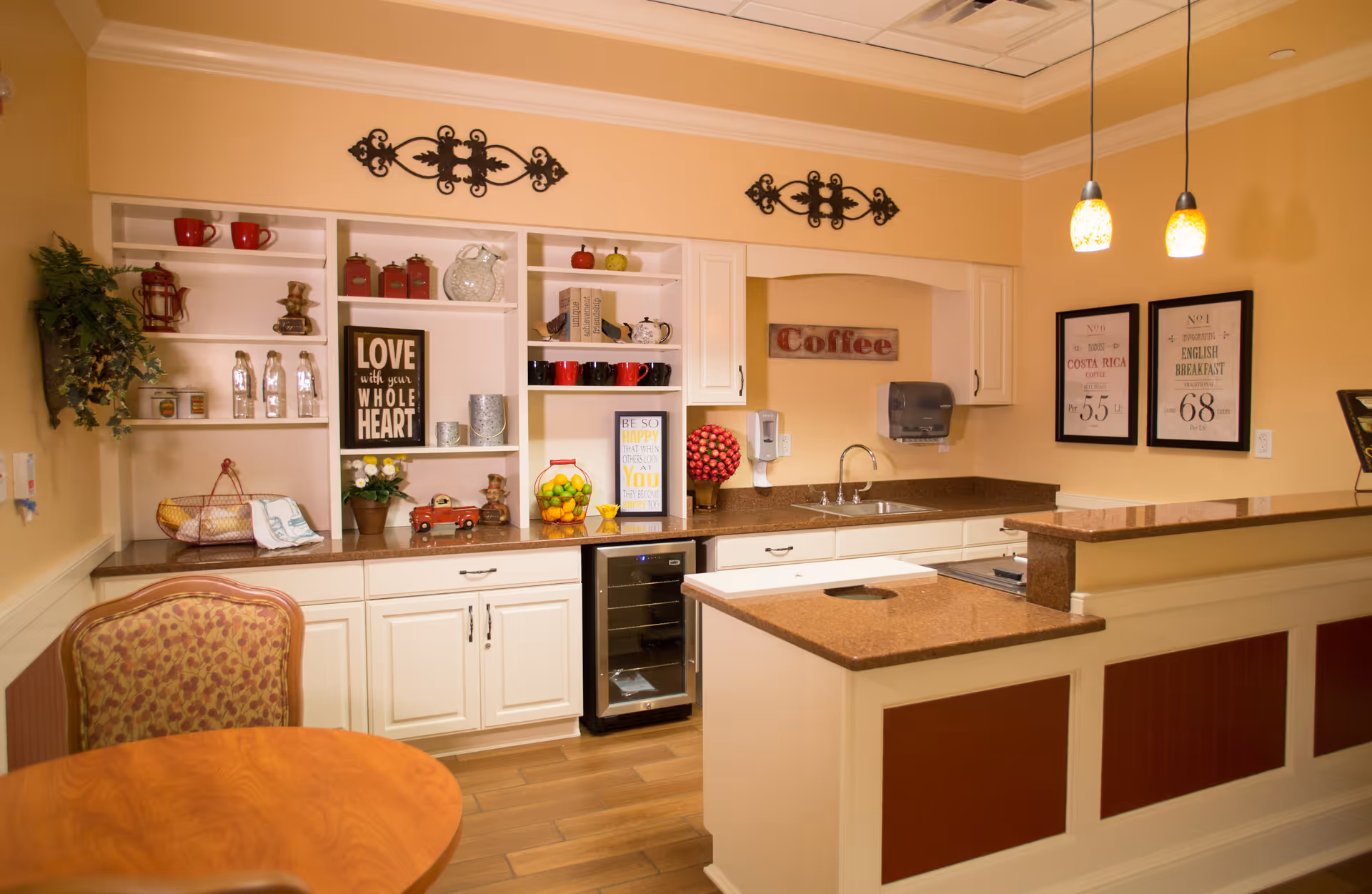 A cozy kitchen area with white cabinets, brown countertops, and wooden flooring. The shelves are decorated with various items including red mugs, glass jars, a basket with fruit, and framed inspirational quotes. There is a small wine cooler under the counter, a sink with a paper towel dispenser above it, and two hanging pendant lights. A round wooden table with a patterned chair is visible in the foreground.