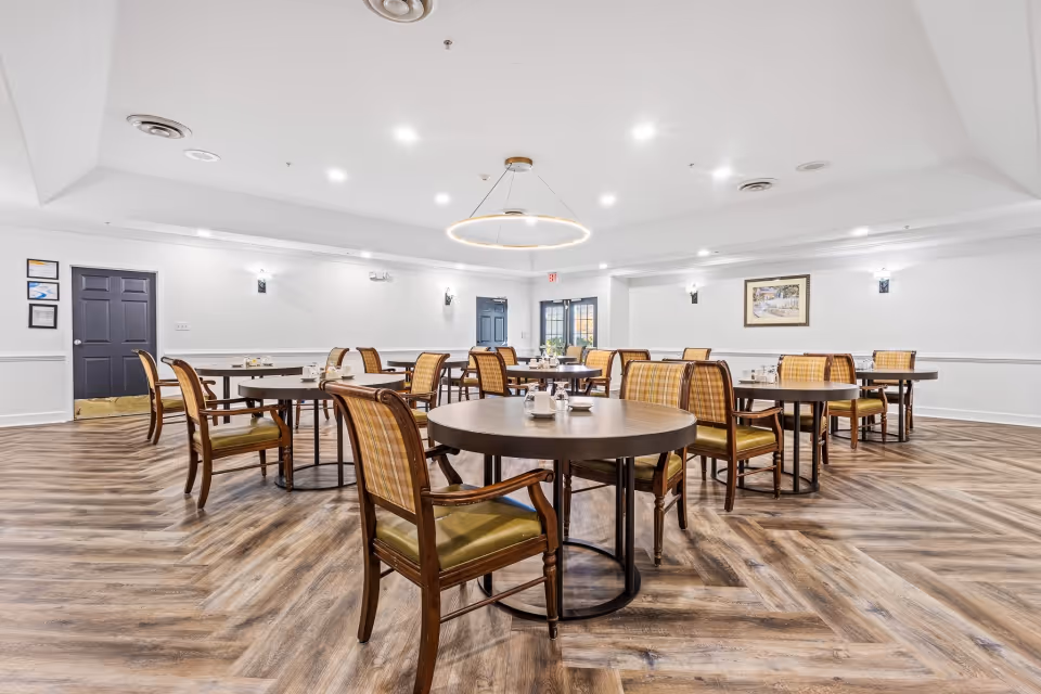 A spacious senior living dining room with multiple round tables and wooden chairs with cushioned seats and backs. The room has wood-patterned flooring, white walls with wainscoting, and a modern circular light fixture hanging from the ceiling. There are framed pictures on the walls and double doors at the far end of the room.