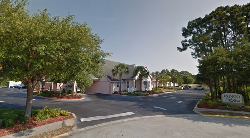 Exterior view of Indigo Palms facility showing a single-story building with a gray roof, surrounded by palm trees and other greenery. There is a parking lot with several cars and a sign that reads 'Indigo Palms' near the entrance.