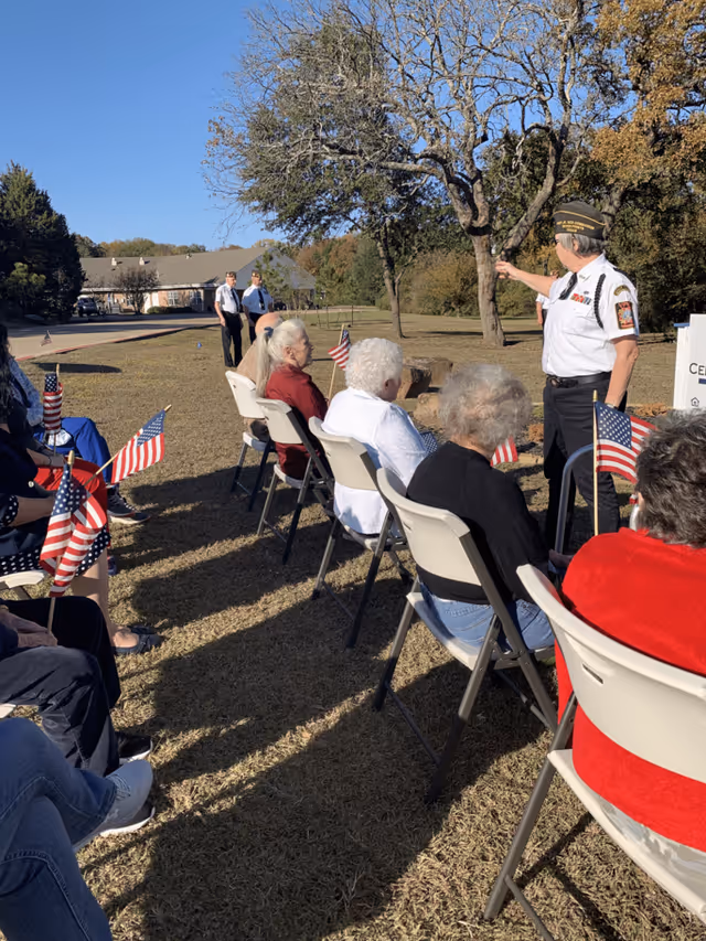 A group of elderly people seated outdoors on folding chairs, holding small American flags. A man in a military uniform stands in front of them, pointing towards something off-camera. The setting is a grassy area with trees and a building in the background under a clear blue sky.