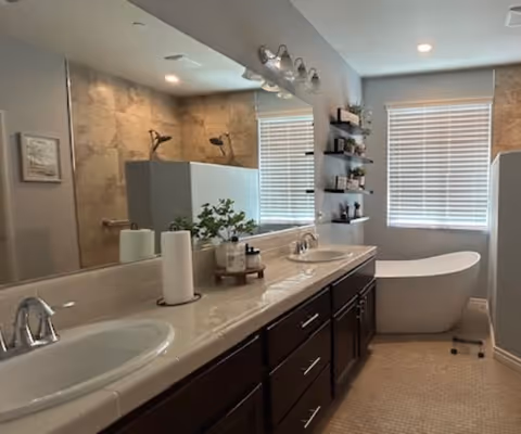 A modern bathroom featuring a double sink vanity with dark wood cabinets and a large mirror above. There is a freestanding white bathtub near a window with blinds, and a tiled shower area with a glass partition. Shelves with decorative items are mounted on the wall next to the bathtub.