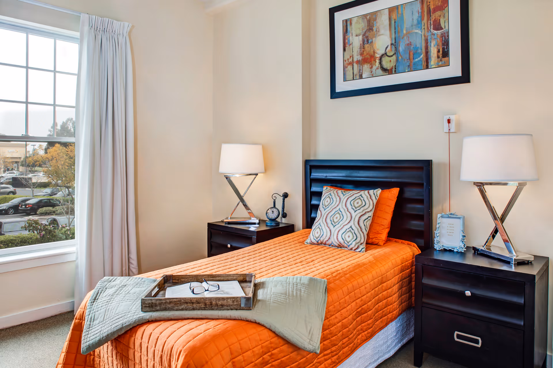 A bright bedroom with a single bed covered in an orange quilt and a decorative pillow. There are two dark wooden nightstands on either side of the bed, each with a modern lamp. A framed abstract painting hangs above the bed. A window with white curtains lets in natural light, showing a parking lot and trees outside. A wooden tray with reading glasses and a book rests on the bed.