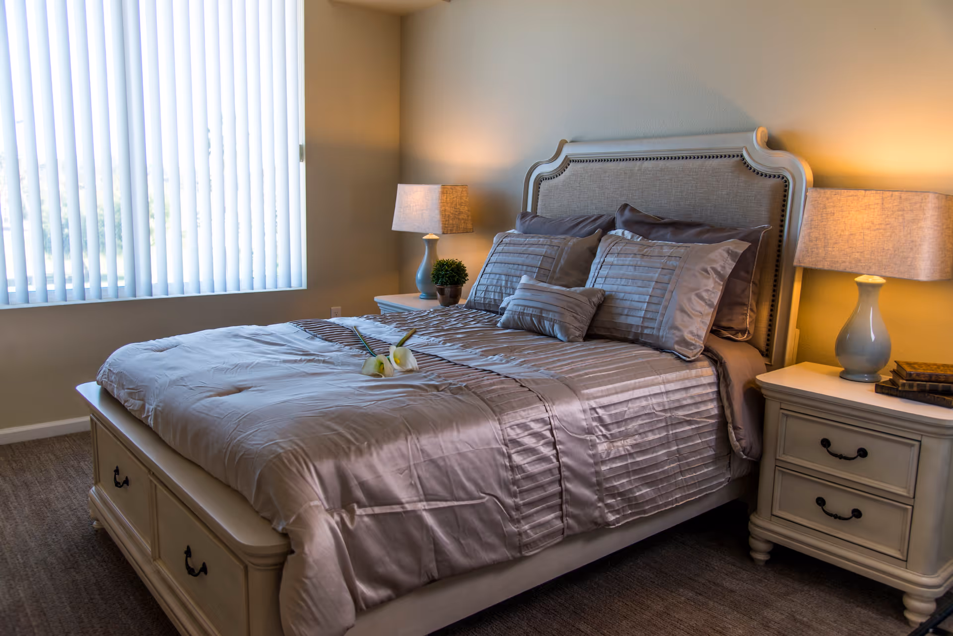 A neatly made bed with a padded headboard in a bedroom. The bed has multiple pillows and a light gray comforter with a decorative runner. There are two bedside tables, each with a lamp and one with a small plant and the other with a stack of books. A large window with vertical blinds allows natural light into the room.