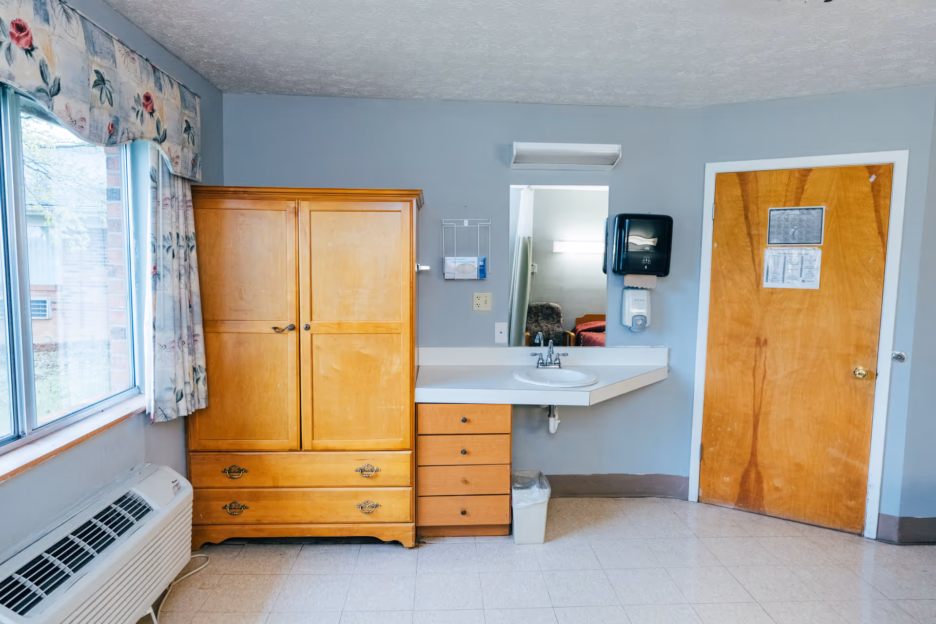 Interior view of a room with a wooden wardrobe, a small set of drawers, a wall-mounted sink with a mirror above it, a soap dispenser, and a wooden door with papers attached. There is a large window with floral curtains and an air conditioning unit below it.