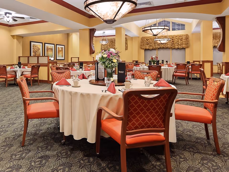Elegant dining room with round tables set with white tablecloths, red upholstered chairs, floral centerpieces, and chandeliers.
