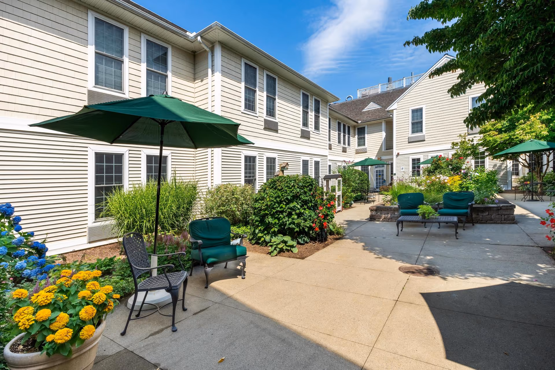 Outdoor courtyard area at East Village Place with beige two-story buildings surrounding a paved patio. The patio features green cushioned chairs, metal chairs, tables with green umbrellas, and various colorful flowers and shrubs along the edges. The sky is clear and blue.
