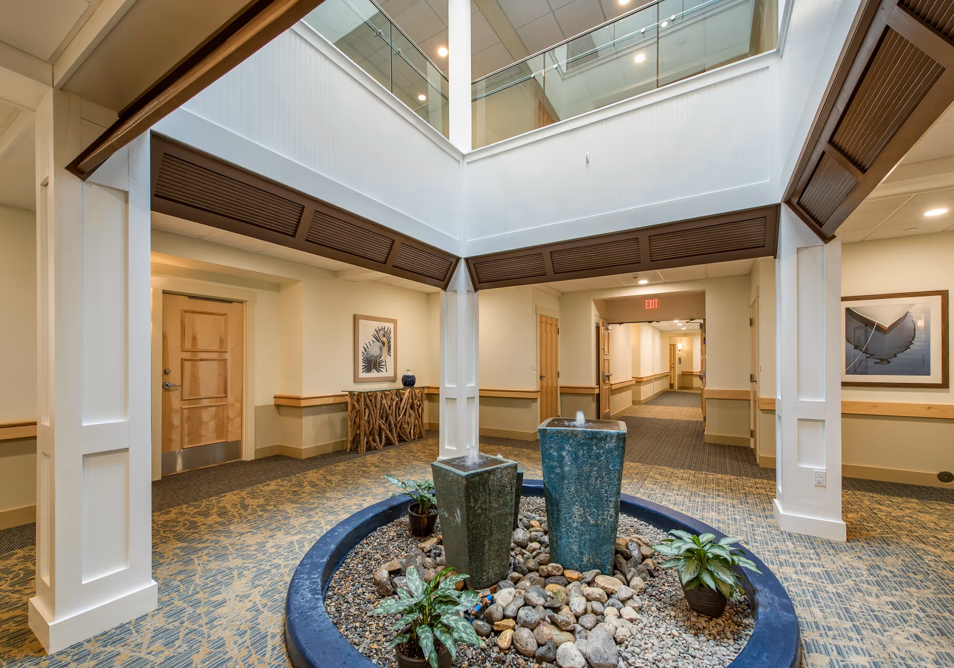 Atrium-style interior hallway featuring a central pebble-filled fountain with two blue stone water features, potted plants, and surrounding corridors and doors.