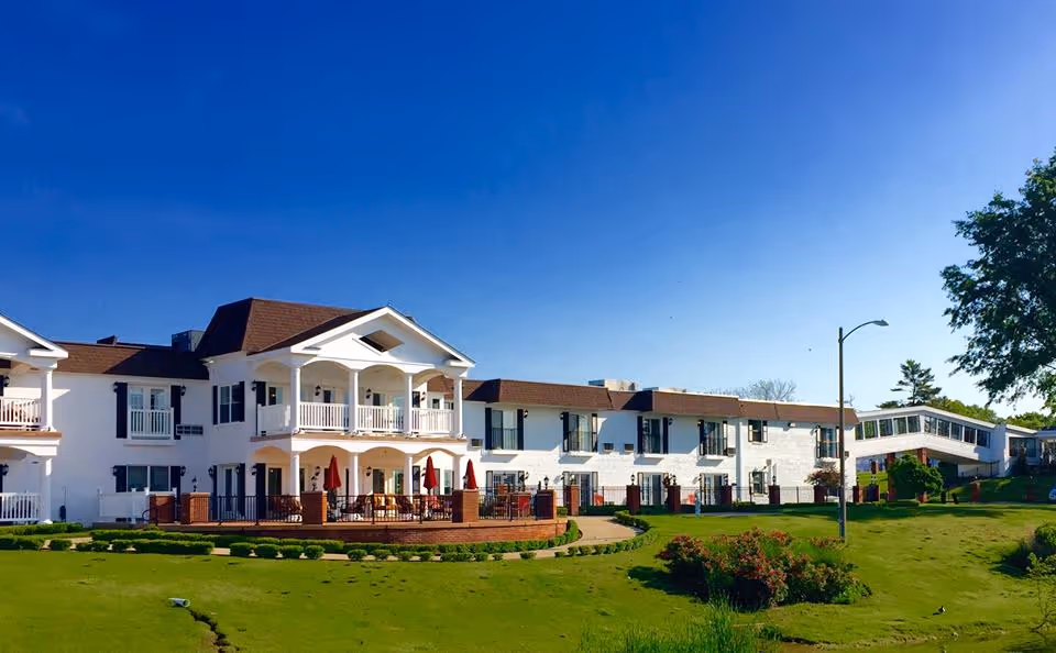Exterior view of a large white two-story nursing home building with a brown roof, balconies, and a patio area with red umbrellas. The building is surrounded by a well-maintained green lawn and some bushes under a clear blue sky.