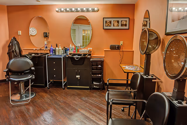 Interior of a salon room with orange walls, featuring two black salon chairs with hair dryers, a black styling chair, mirrors on the wall, and various hair care products on counters.