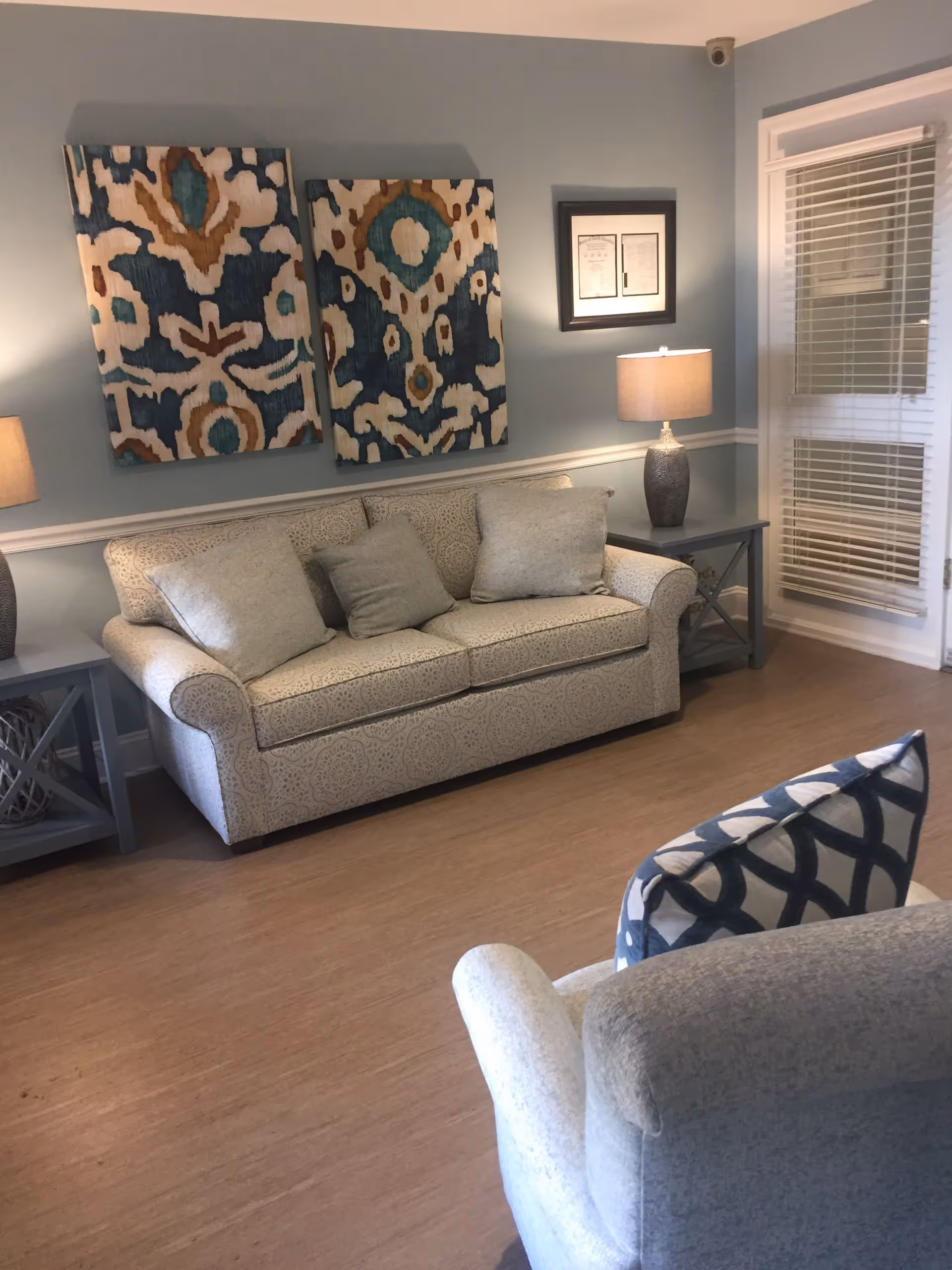 A cozy living room with a patterned beige sofa adorned with three cushions, a matching armchair with a blue and white patterned pillow, two side tables each with a lamp, two abstract paintings on the wall, and a window with blinds.