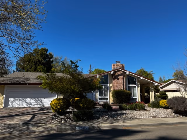 Exterior view of a single-story residential building with a brick chimney, large windows, and a two-car garage. The front yard features various shrubs, small trees, and decorative rocks under a clear blue sky.