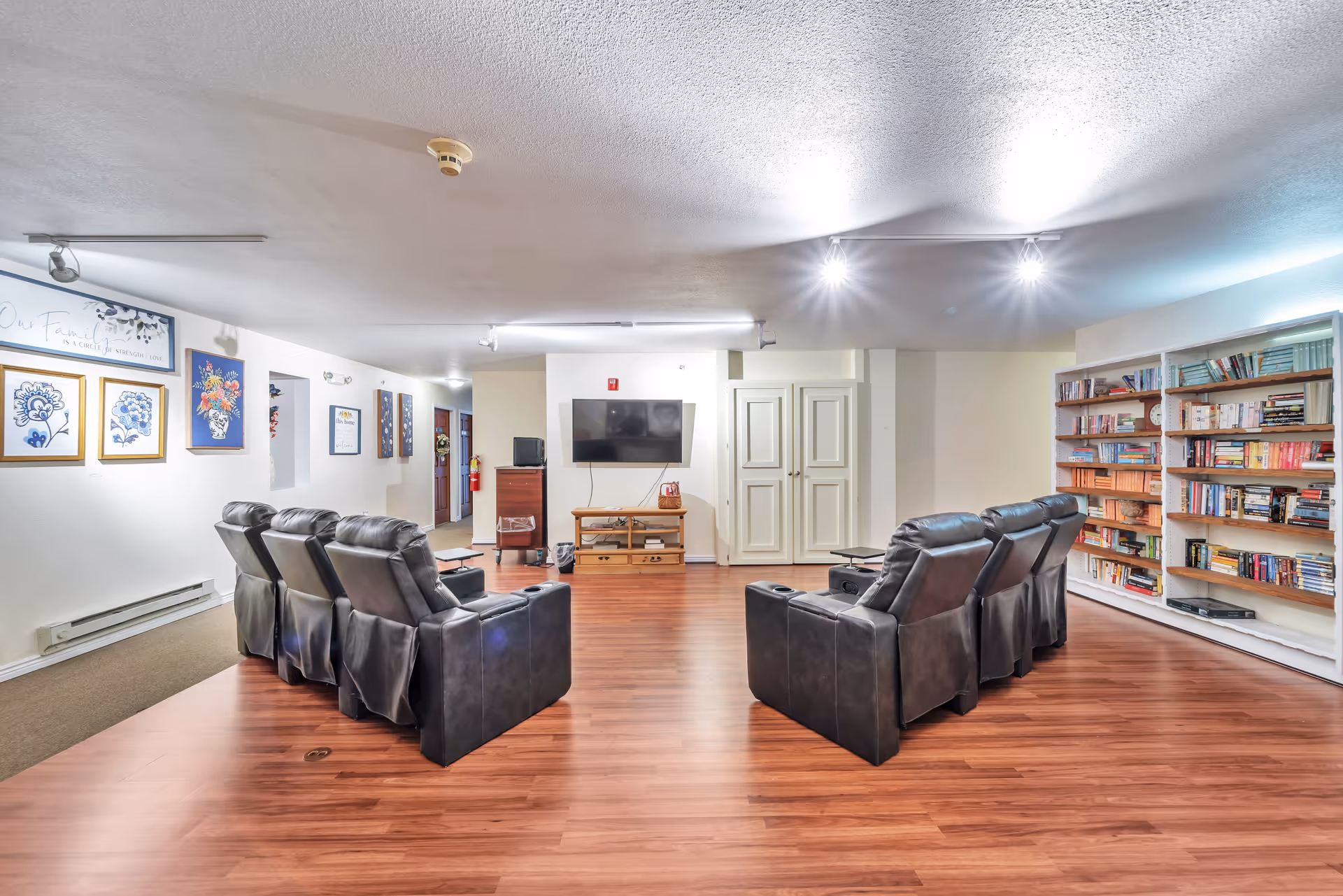 A senior living facility common area with two rows of black leather recliners facing a wall-mounted flat screen TV. The room has wooden flooring, white walls with framed artwork, and a large bookshelf filled with books on the right side. The ceiling has multiple light fixtures providing bright illumination.