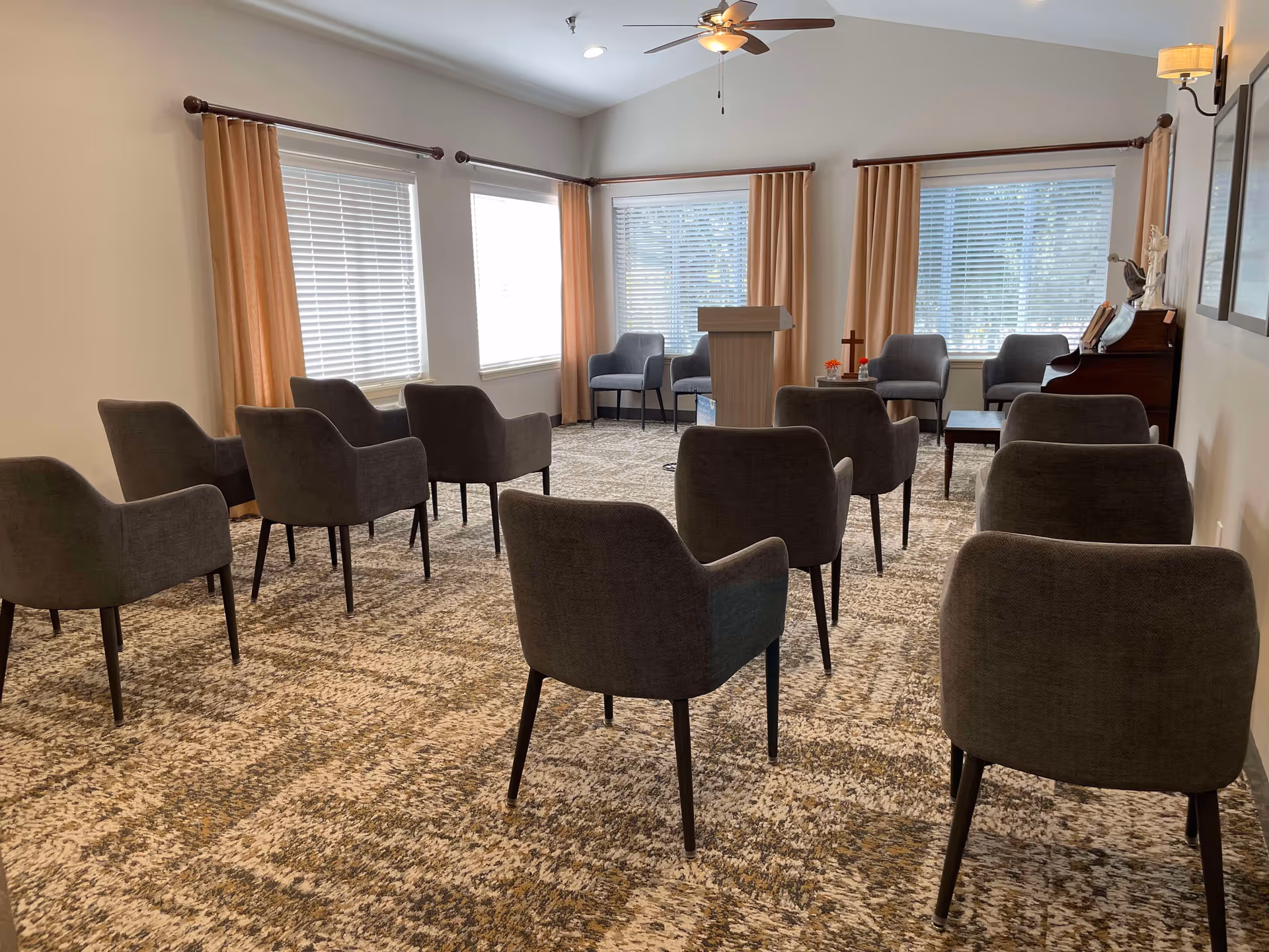 A small meeting or chapel room with multiple gray upholstered chairs arranged in rows facing a wooden podium. The room has large windows with beige curtains, a ceiling fan with a light, a piano on the right side, and a carpeted floor with a patterned design.
