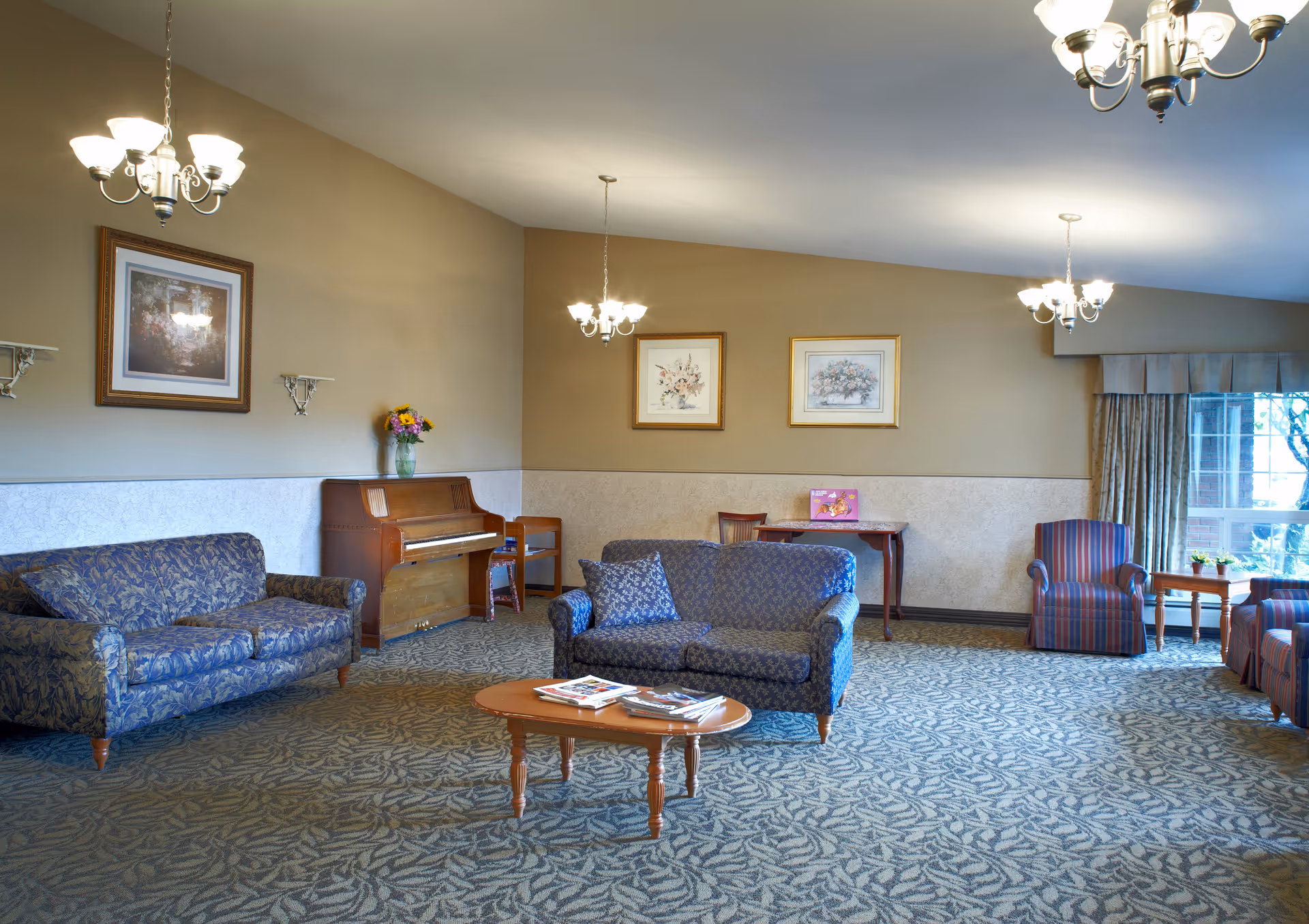 A spacious living room area with patterned carpet, two blue floral sofas, two striped armchairs, a wooden coffee table with magazines, a wooden piano with a vase of flowers on top, framed floral artwork on the walls, and multiple chandeliers providing warm lighting. A window with curtains allows natural light to enter the room.