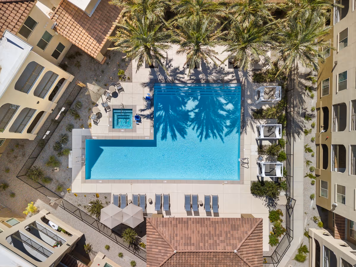 Aerial view of a rectangular swimming pool with clear blue water surrounded by palm trees and lounge chairs. There is a smaller hot tub adjacent to the pool with chairs around it. The pool area is enclosed by buildings with beige walls and tiled roofs, and there are shaded cabanas on one side.