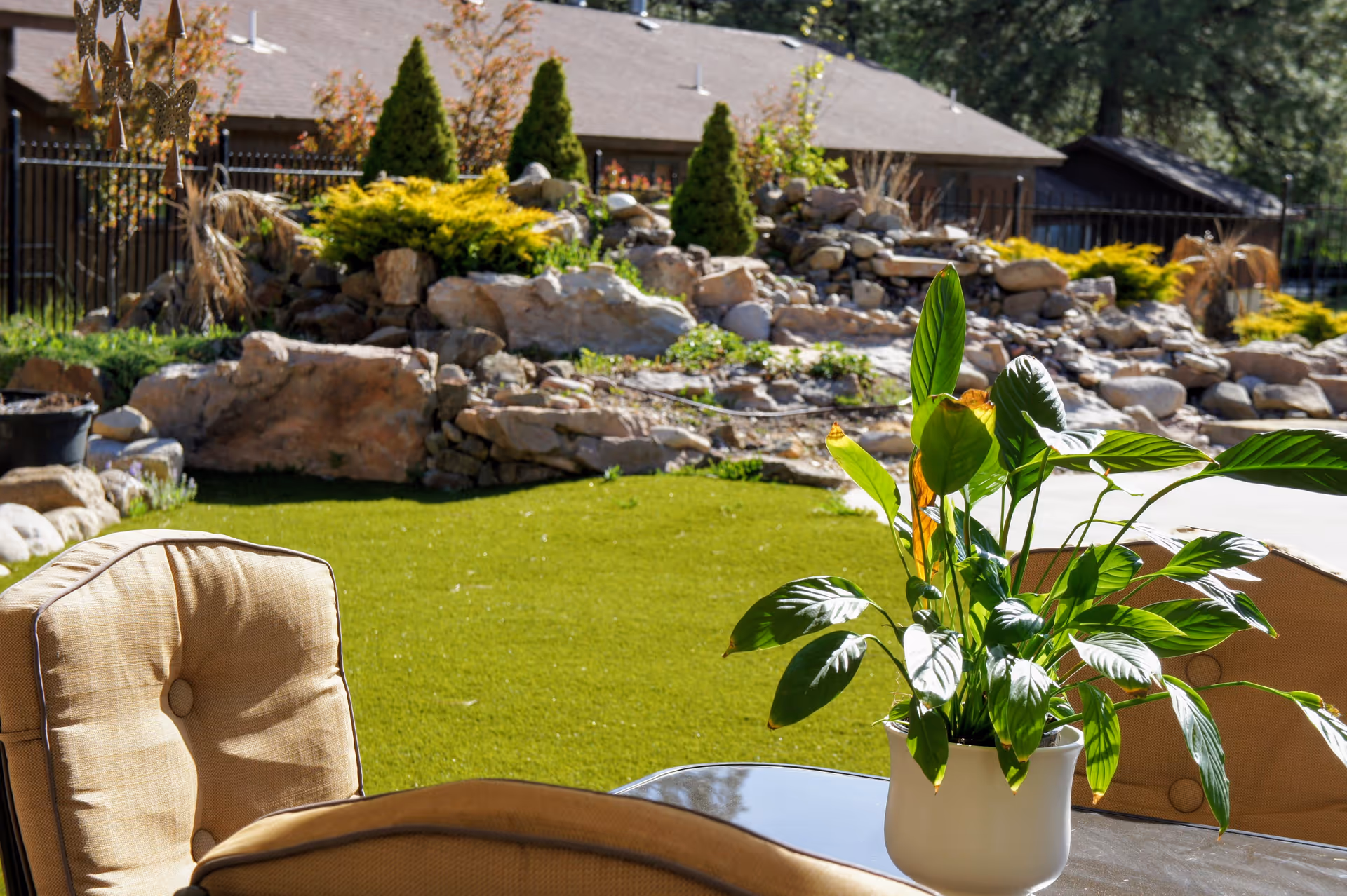 Sunlit patio with cushioned chairs and a potted green plant overlooking a landscaped rock garden and lawn.