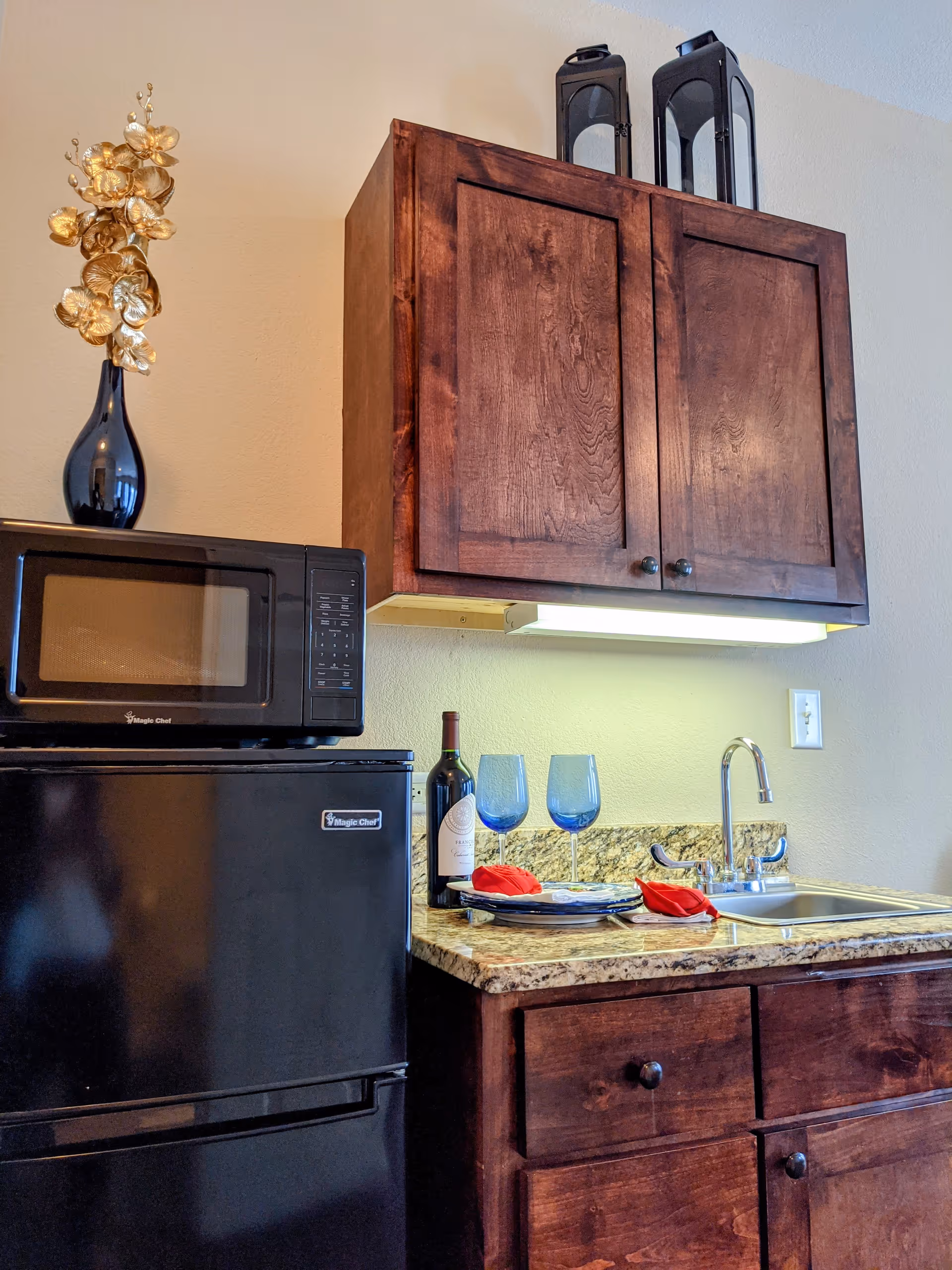 A small kitchen area with dark wooden cabinets, a granite countertop, a stainless steel sink with a faucet, a black Magic Chef microwave on top of a black Magic Chef mini refrigerator. On the countertop, there is a bottle of wine, two blue wine glasses, and two plates with red napkins. A decorative black vase with golden flowers is placed on top of the microwave, and two black lanterns are on top of the upper cabinet.