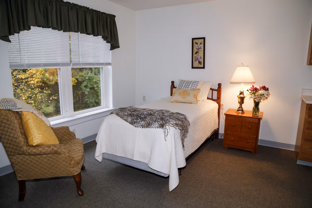 A cozy bedroom with a single bed covered in a white quilt and decorative pillows. A knitted throw is draped over the foot of the bed. Next to the bed is a wooden nightstand with a lamp and a vase of flowers. A cushioned armchair with a yellow pillow is placed near a window with white blinds and a dark green valance. The walls are white, and there is a framed floral picture hanging above the bed.