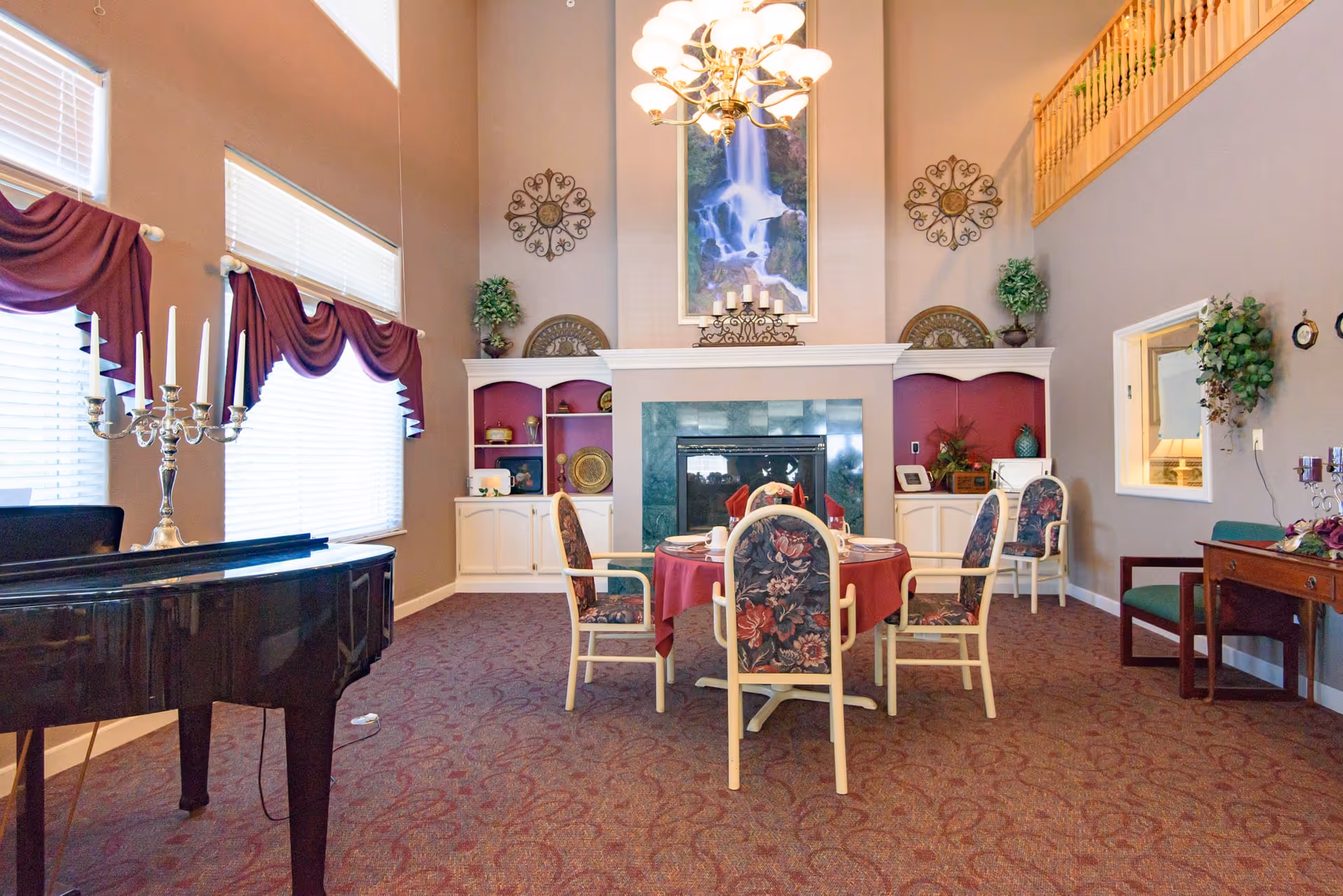 A cozy dining area in a senior living facility with a round table covered with a red tablecloth and set with cups and napkins. The room features floral upholstered chairs, a fireplace with a green tile surround, built-in white cabinets with decorative items, a large painting of a waterfall above the fireplace, and a chandelier hanging from the high ceiling. There is a black grand piano with a silver candelabra on it near large windows with white blinds and purple valances. The carpet is patterned in shades of red and purple, and there are plants and wall decorations adding to the warm ambiance.
