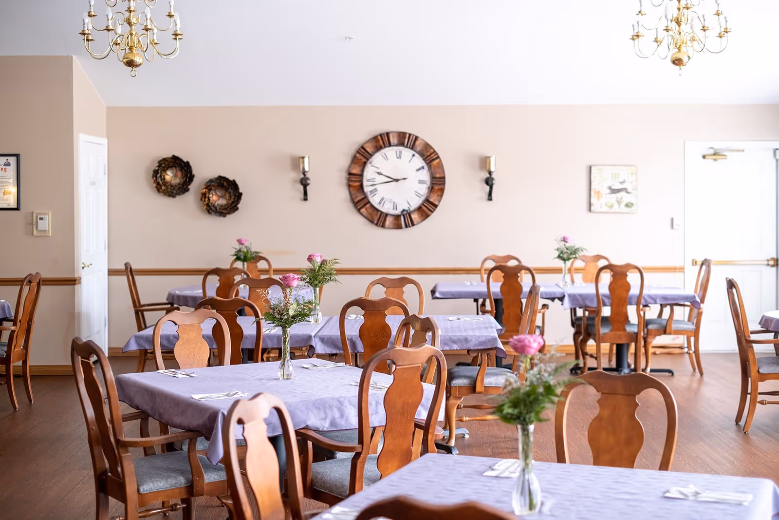 A dining room with several wooden tables covered with lavender tablecloths, each set with silverware and a small vase containing pink flowers and greenery. The room has wooden chairs with cushioned seats, beige walls adorned with a large round clock, two decorative wall sconces, and two wreaths. Two chandeliers hang from the ceiling, and there is a white door on the right side of the room.