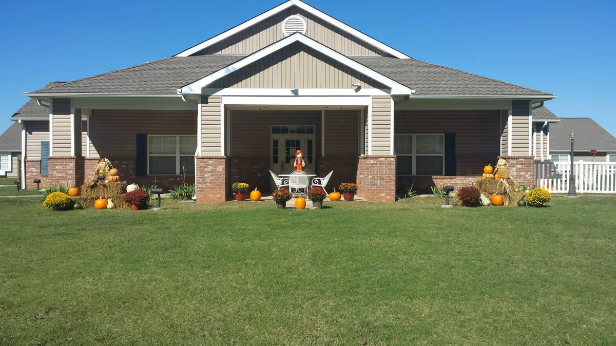 Front view of a single-story assisted living building with a wide lawn and autumn decorations including pumpkins and hay bales.