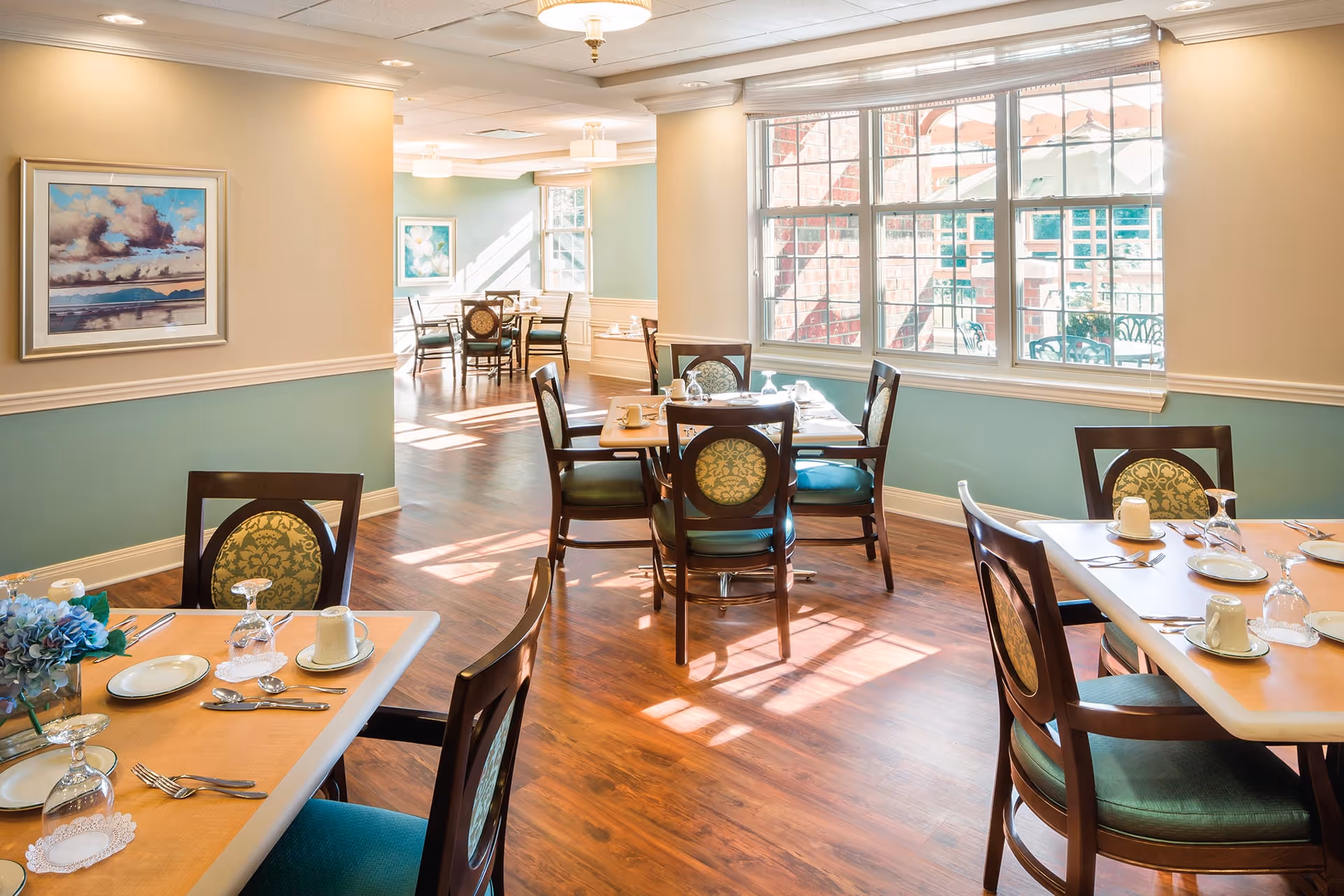A bright dining room with wooden floors and several tables set with plates, cups, and silverware. The room has large windows letting in natural light, light blue and beige walls, and framed artwork. Chairs with patterned upholstery surround the tables.