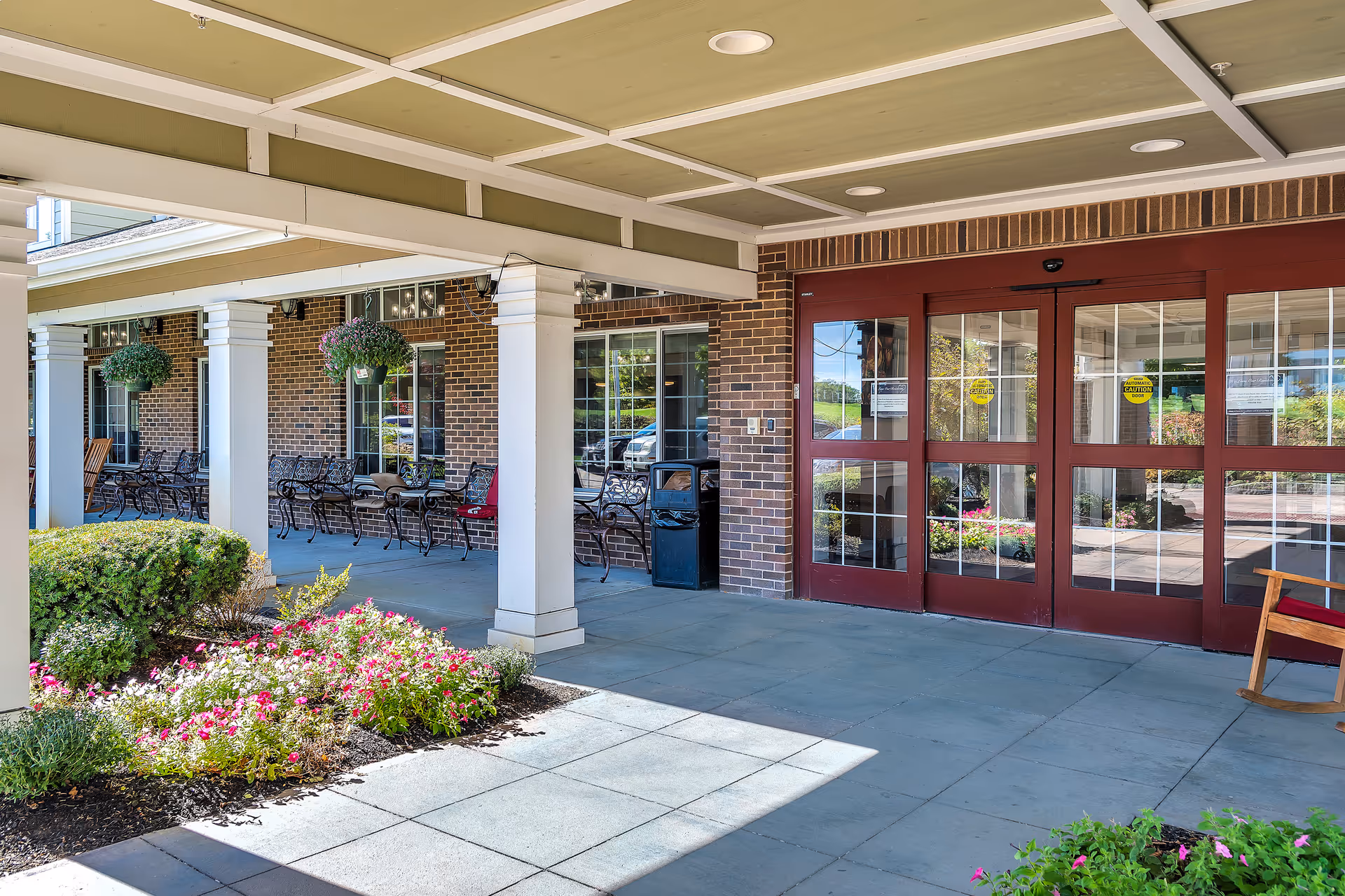 Covered entrance and patio with seating, flower beds, and glass entry doors of a retirement community.