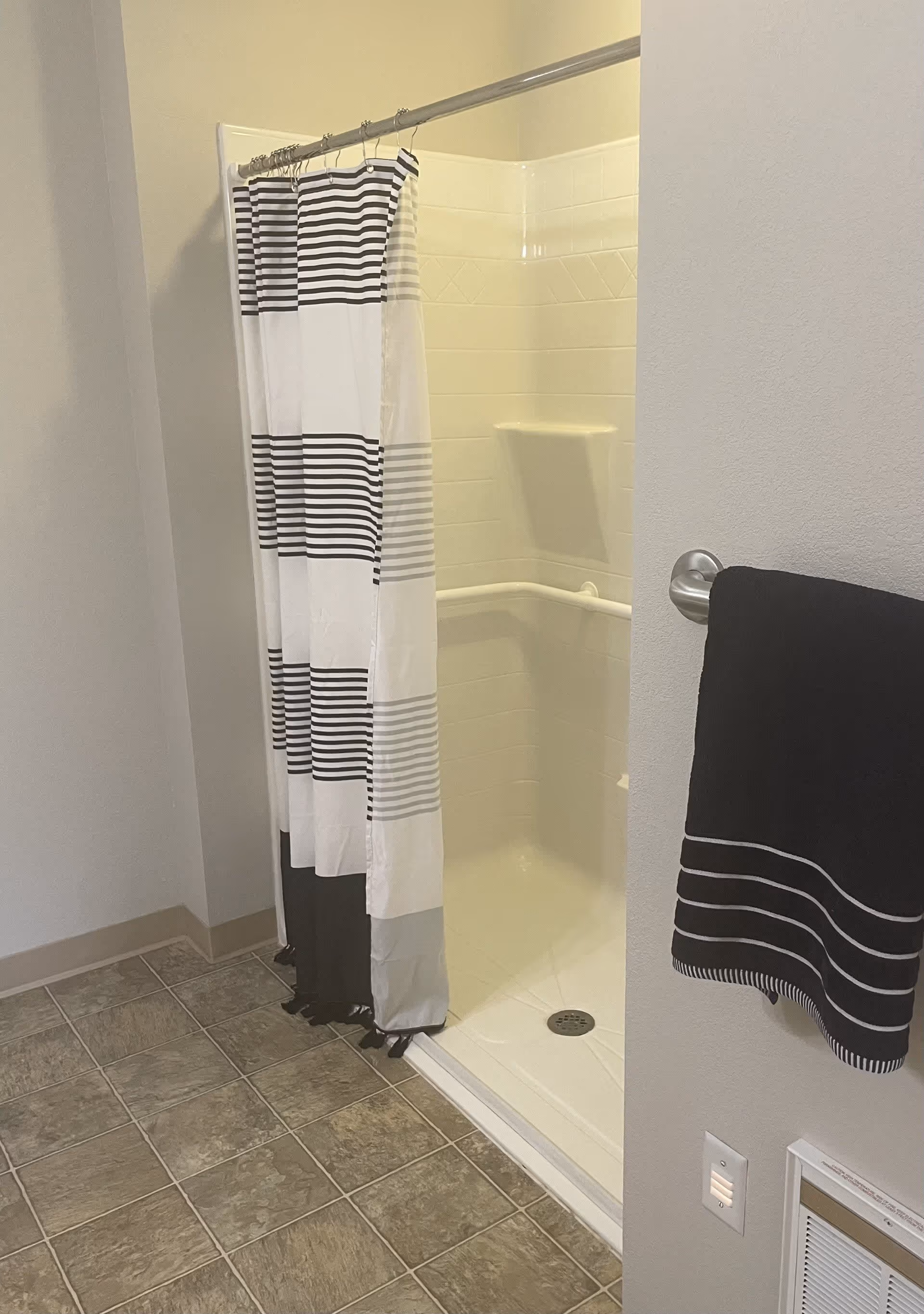 A bathroom shower area with a white and black striped shower curtain partially drawn. The shower has white tiled walls with a built-in corner shelf and a grab bar. A black towel with white stripes hangs on a silver towel rack mounted on the wall next to the shower. The floor is covered with grayish-brown tiles.