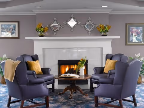 A cozy living room area with four dark blue armchairs arranged around a round wooden coffee table. The table has an open book and a vase with flowers. Behind the chairs is a white fireplace with a fire burning. Above the fireplace are three decorative mirrors and two flower arrangements on either side. The walls are painted gray and have framed artwork. The floor is covered with a patterned blue carpet.