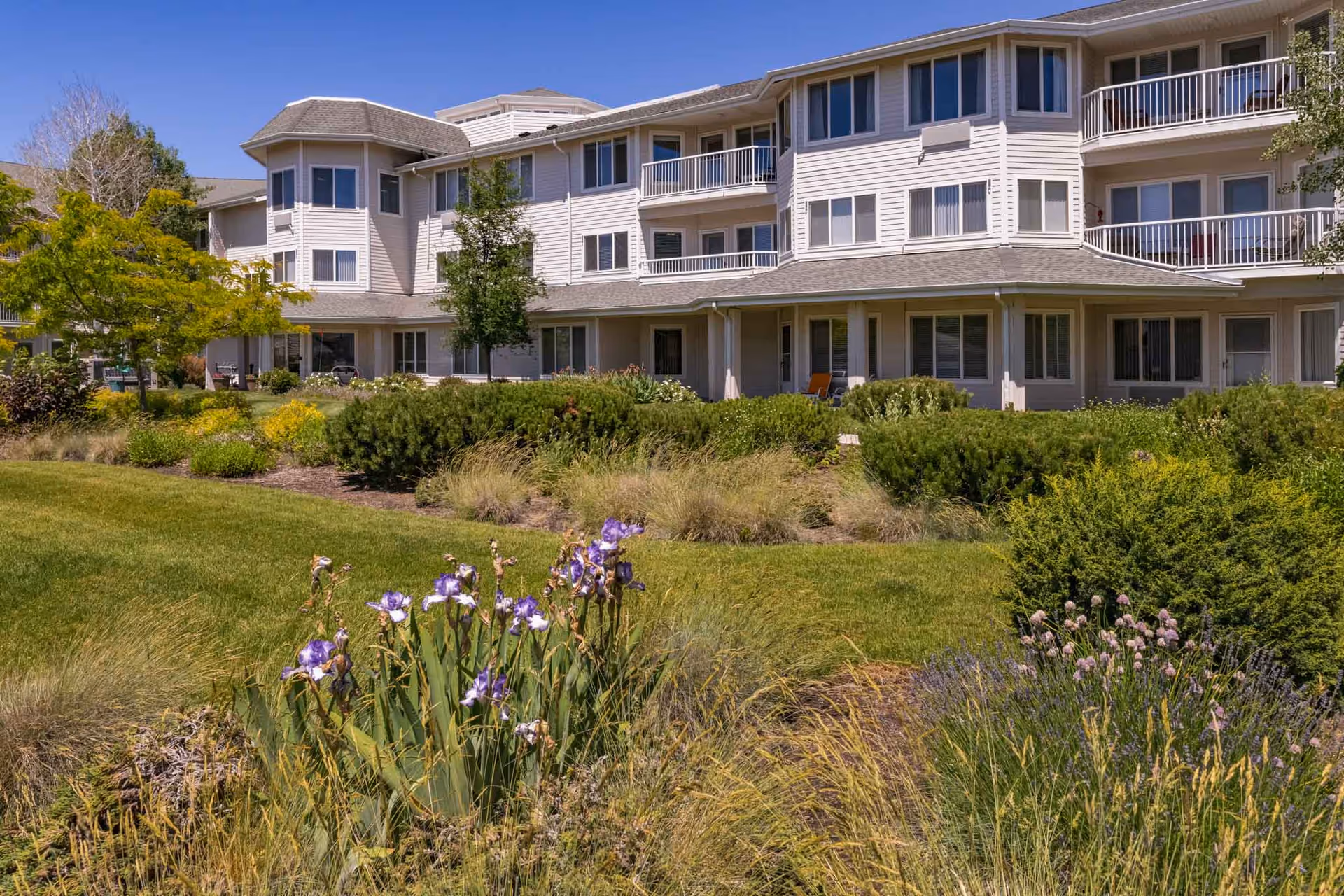Exterior view of a multi-story senior living facility building with balconies and large windows, surrounded by well-maintained gardens featuring green shrubs, flowering plants, and a manicured lawn under a clear blue sky.