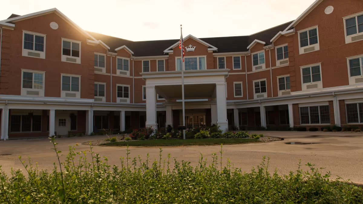 Front entrance of a three-story brick senior living building with a covered porte-cochère, flagpole, and landscaped circular driveway.