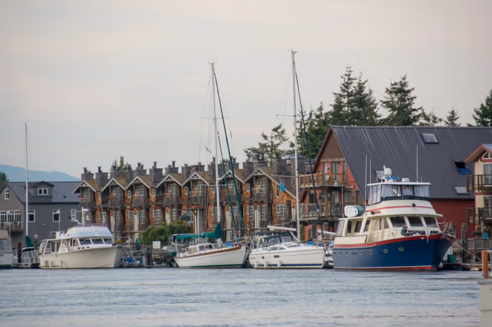 A waterfront scene showing several boats docked in front of a row of multi-story buildings with peaked roofs and chimneys, surrounded by trees and hills in the background.