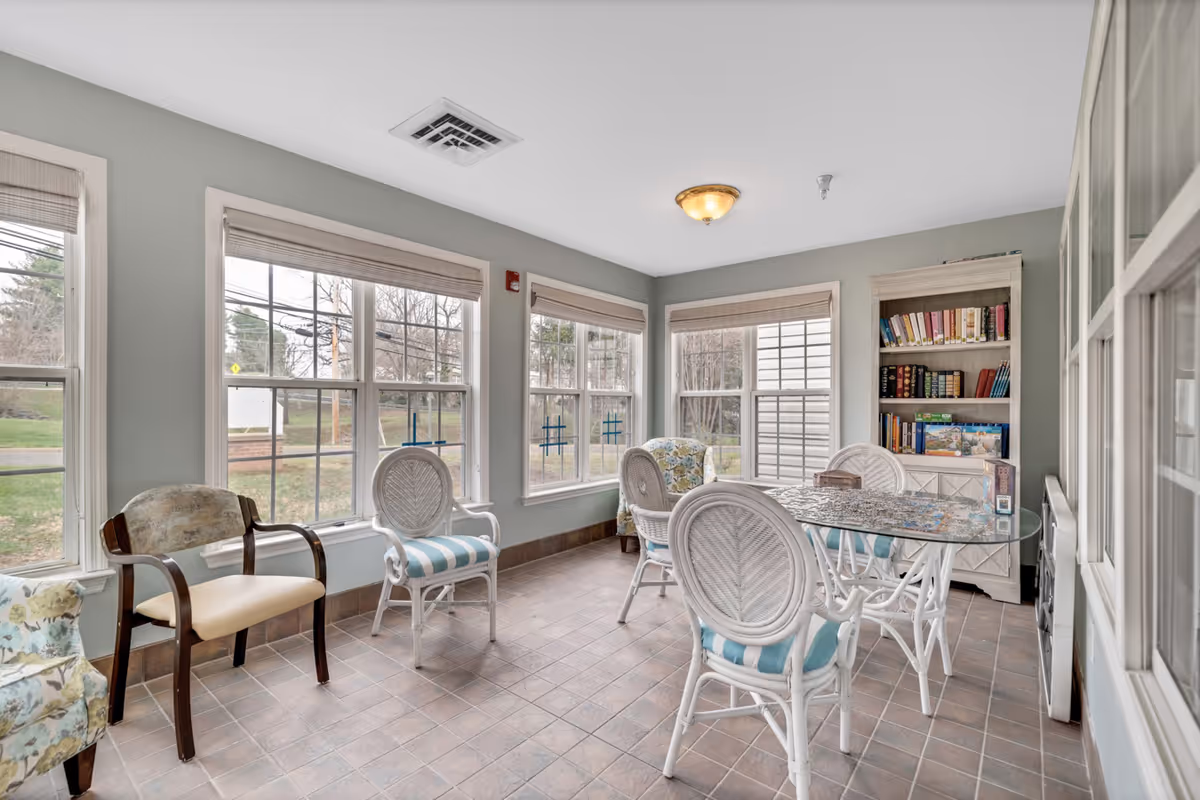 Bright sunroom-style common area with wicker chairs around a glass table, extra seating and a bookshelf next to large windows.