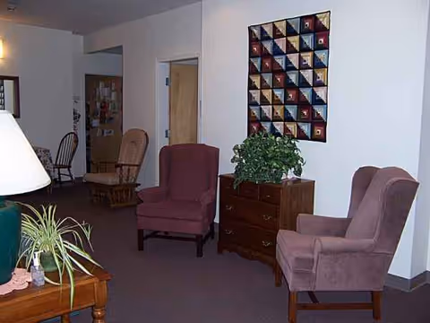 Cozy senior living lounge with pink armchairs, a wooden dresser, and a colorful wall quilt.