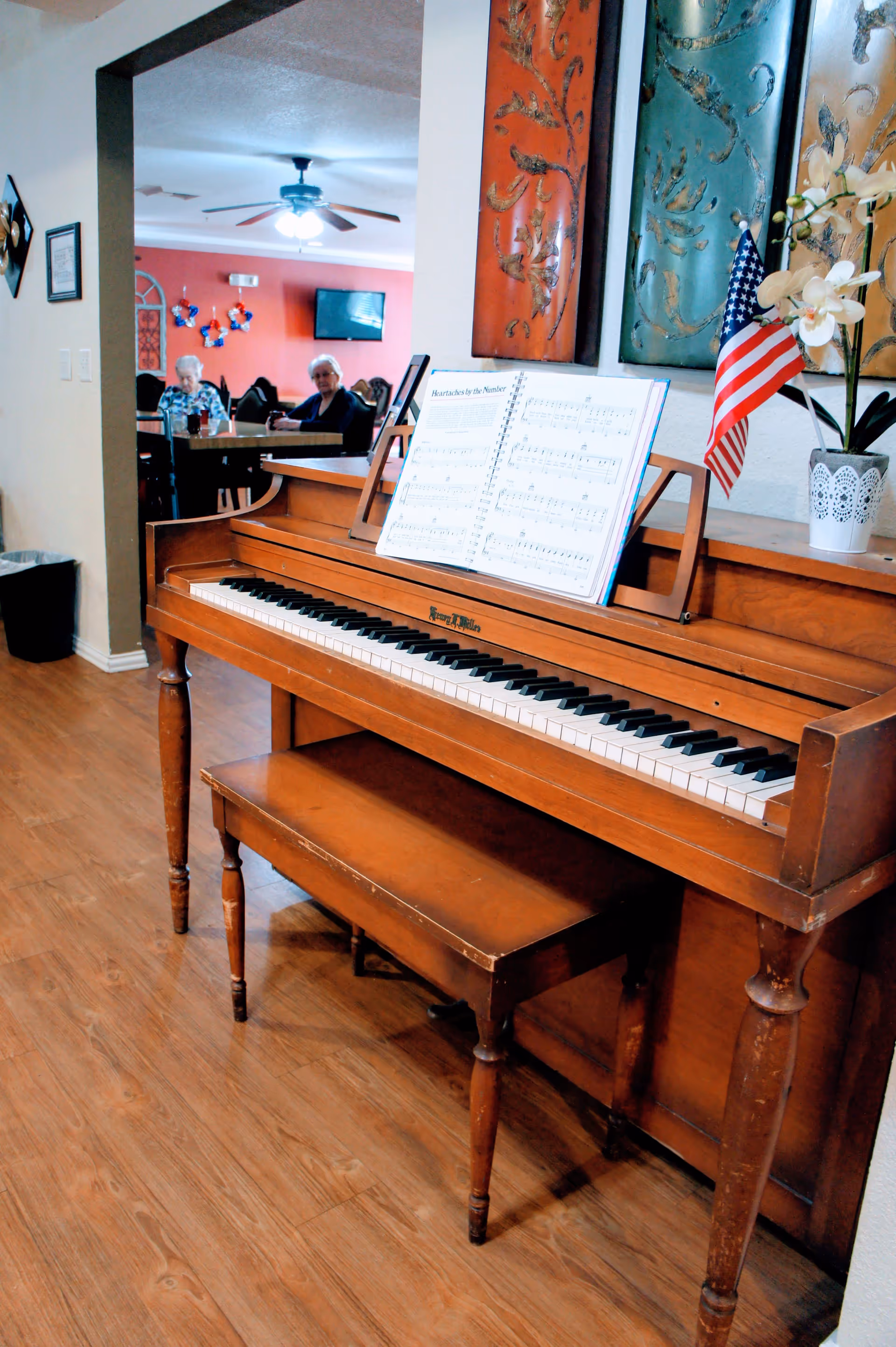 A wooden piano with a matching bench is placed against a wall decorated with three colorful metal art panels and a small American flag in a white pot with flowers. In the background, two elderly women are seated at a table in a room with red walls, a ceiling fan, and a wall-mounted TV.