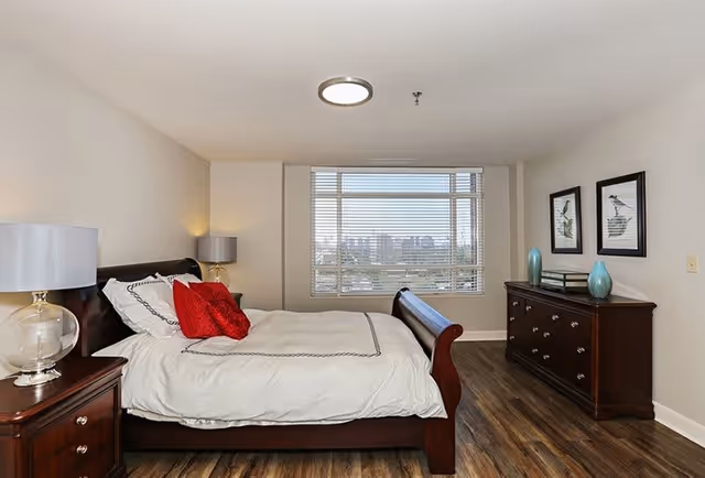 A bedroom with a large wooden bed featuring white bedding and red accent pillows. There are two bedside tables with lamps, a wooden dresser with decorative vases and framed bird artwork on the wall. A large window with blinds offers a view of a cityscape outside.
