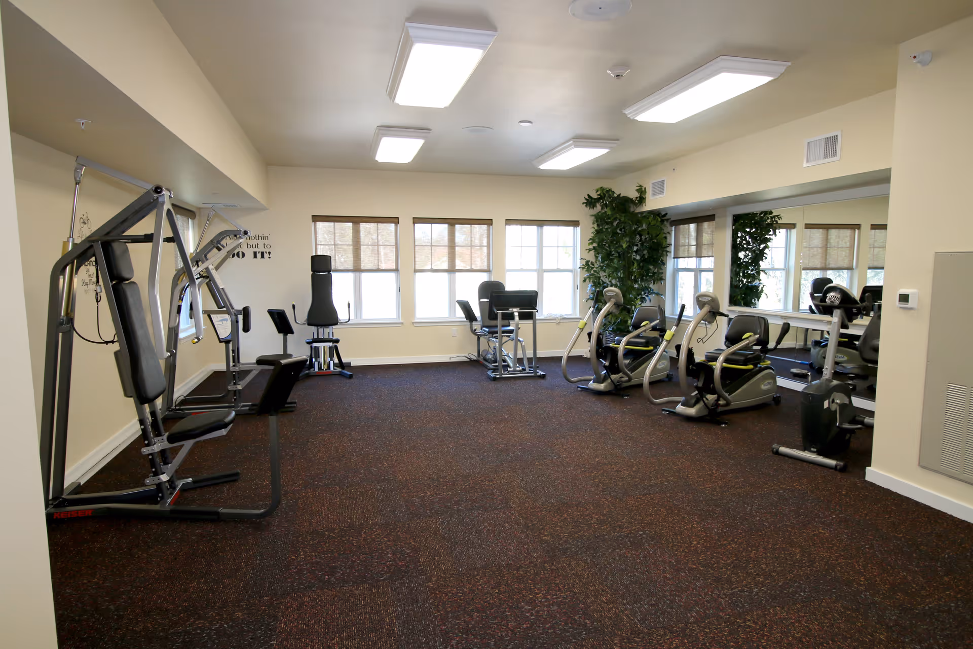 A well-lit exercise room with various fitness equipment including a multi-station gym machine, recumbent bikes, and other workout machines. The room has large windows with blinds, a large mirror on one wall, and a potted plant in the corner.