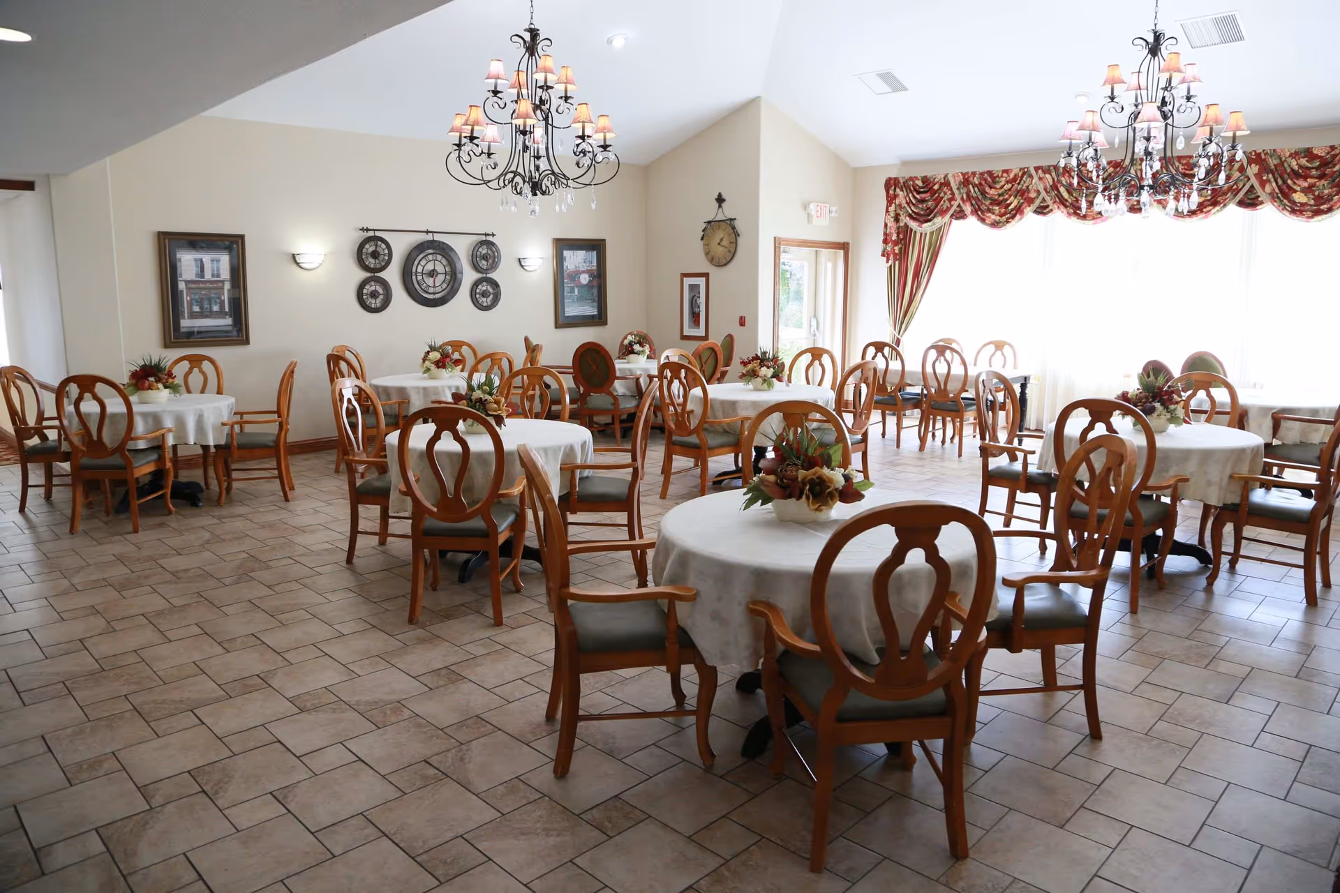 Bright dining room with round tables draped in white tablecloths, wooden chairs, chandeliers, and floral centerpieces.