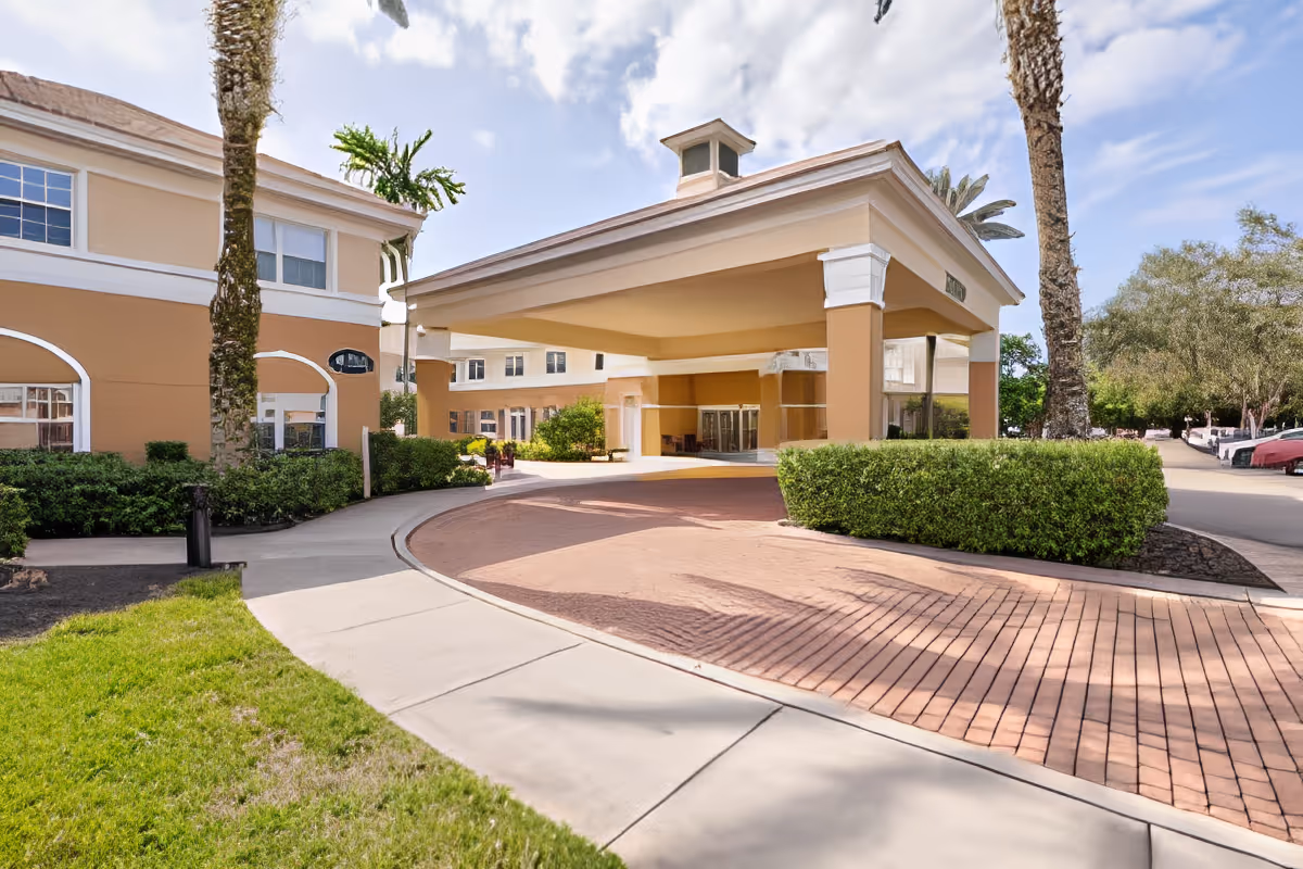 Exterior view of Aston Gardens At Pelican Marsh showing the entrance with a covered driveway, beige and white building facade, palm trees, bushes, and a clear sky.