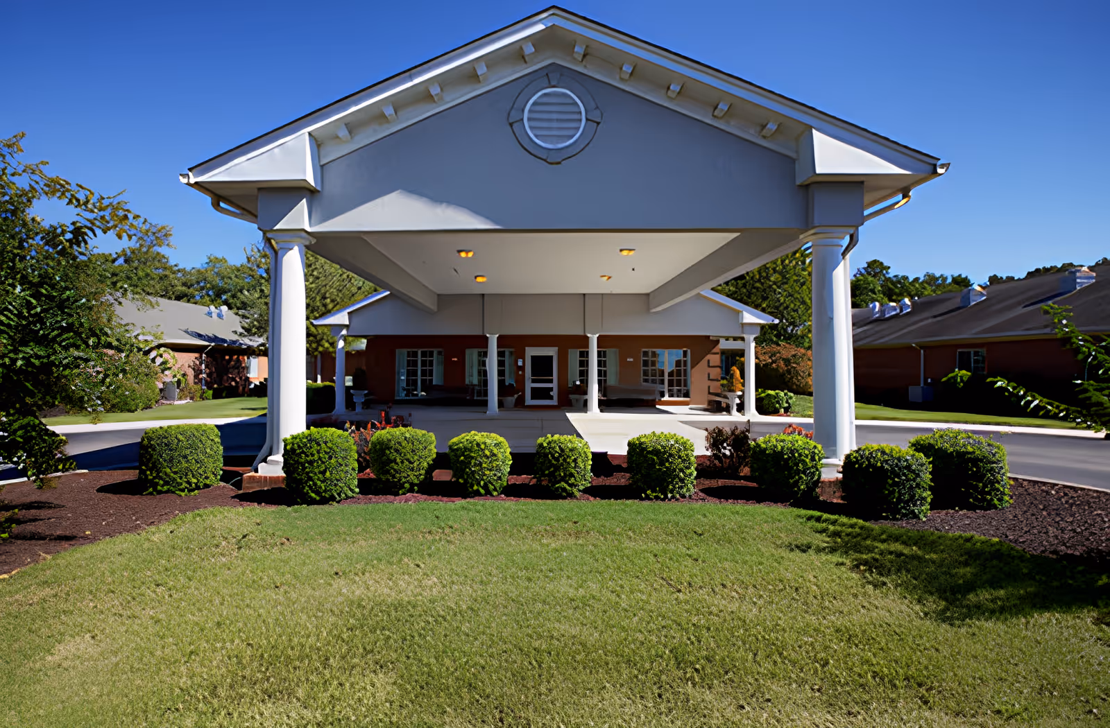 Front exterior view of a senior living facility with a covered entrance supported by white columns, neatly trimmed bushes, green lawn, and clear blue sky.