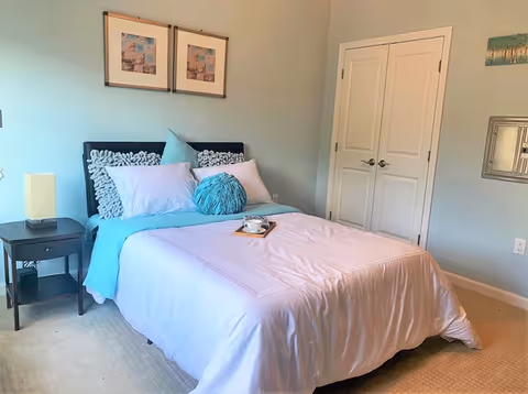 A tidy bedroom with a made bed dressed in pastel bedding, decorative pillows, a nightstand, wall art, and double closet doors.