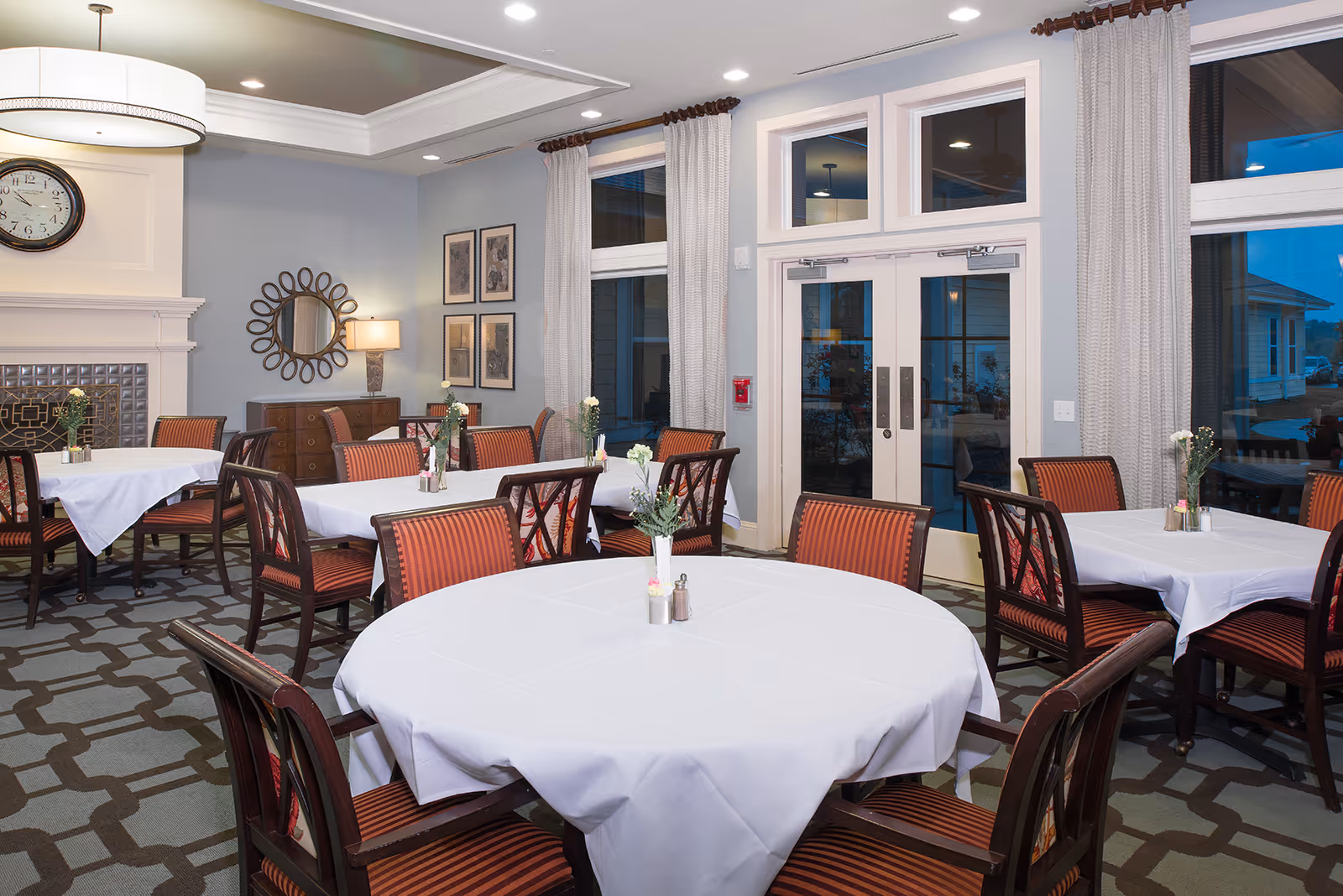 Bright dining room with round and rectangular tables covered in white tablecloths, striped chairs, a decorative wall mirror, and double glass doors.